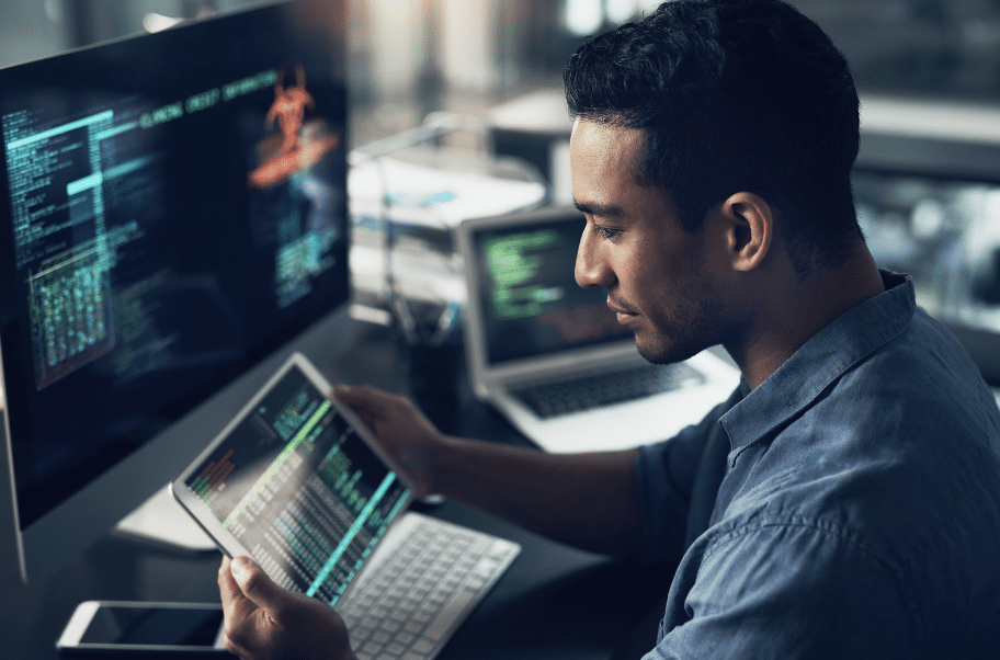 Man coding on a tablet, surrounded by monitors displaying programming code in an office